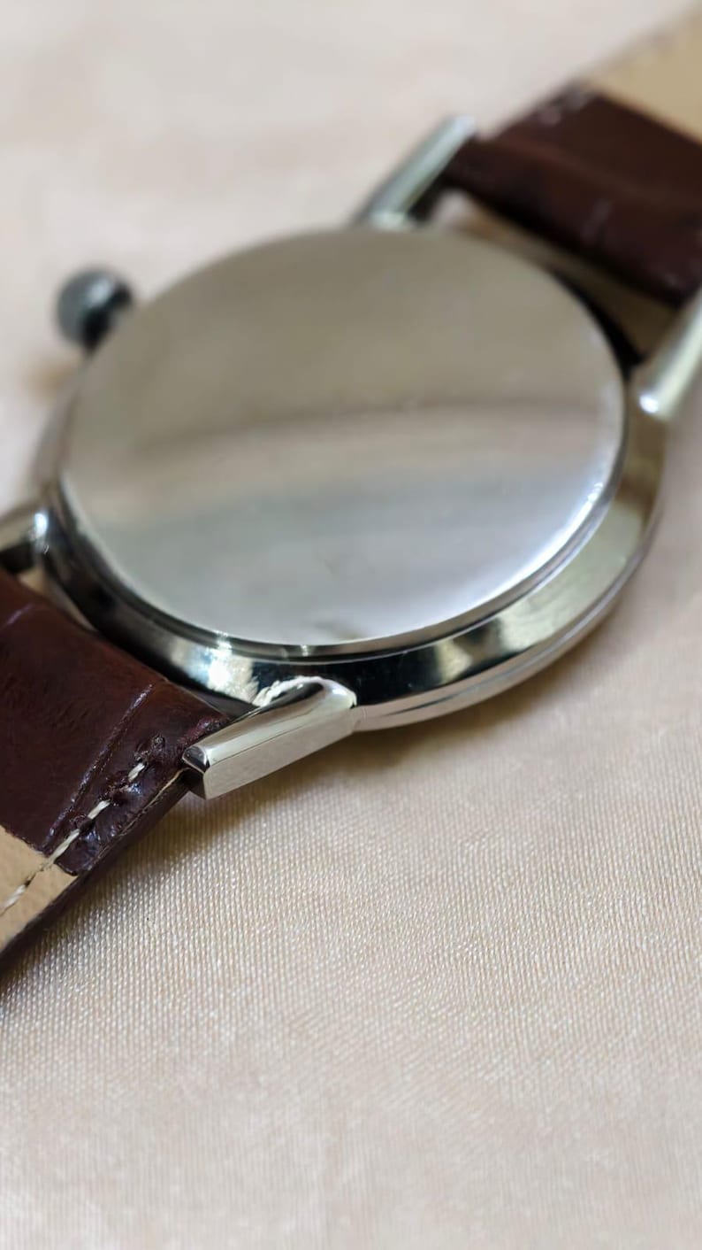 May include: Close-up of a silver wristwatch with a brown leather strap. The watch face is not visible, but the polished metal back and the strap's texture are prominent. The watch's lugs are visible.