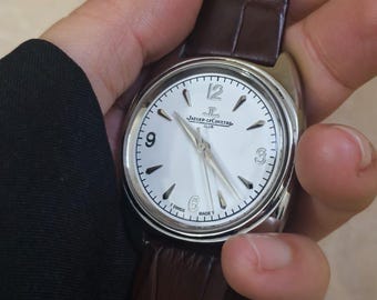 Può includere: Un orologio argentato con quadrante bianco e cinturino in pelle marrone. Il quadrante presenta numeri neri e lancette argentate. Il cinturino è marrone scuro con un motivo strutturato. L'orologio è tenuto in mano.