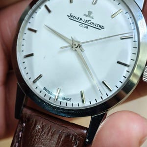 May include: A silver-toned Jaeger-LeCoultre Club watch with a white face and a brown leather strap. The watch face features silver hands and markers, with the brand name and "SWISS MADE" text. The strap is a rich brown color.