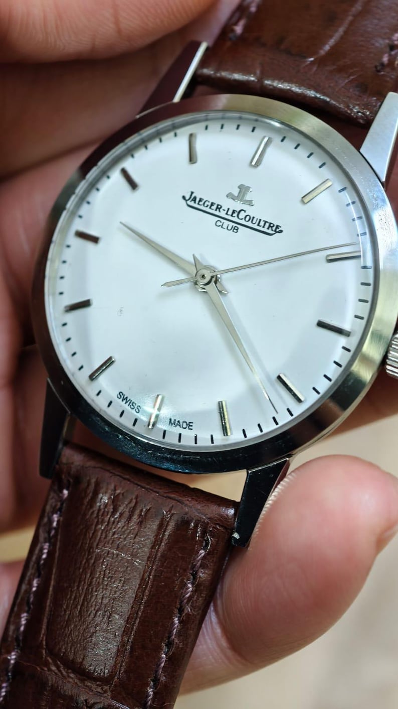 May include: A silver-toned Jaeger-LeCoultre Club watch with a white face and a brown leather strap. The watch face features silver hands and markers, with the brand name and "SWISS MADE" text. The strap is a rich brown color.