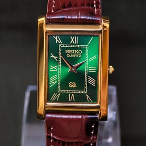 May include: A rectangular gold-toned Seiko Quartz watch with a green face and brown leather band. The watch face features Roman numerals and the Seiko Quartz logo. The watch band is a dark brown, textured leather.