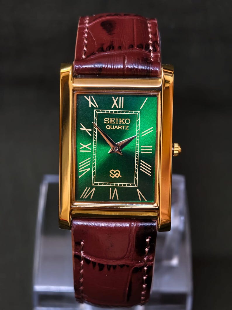 May include: A rectangular gold-toned Seiko Quartz watch with a green face and brown leather band. The watch face features Roman numerals and the Seiko Quartz logo. The watch band is a dark brown, textured leather.