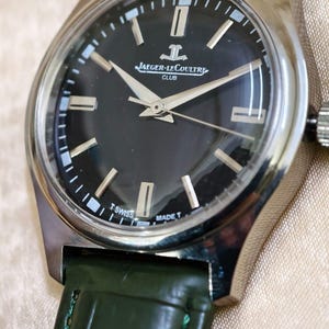 May include: A silver-toned Jaeger-LeCoultre Club watch with a black dial and silver hands. The watch has a green leather strap. The dial features the Jaeger-LeCoultre logo and the words "SWISS MADE".