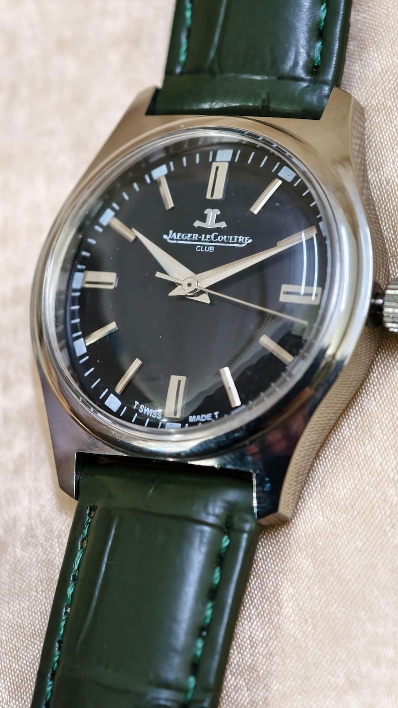 May include: A silver-toned Jaeger-LeCoultre Club watch with a black dial and silver hands. The watch has a green leather strap. The dial features the Jaeger-LeCoultre logo and the words "SWISS MADE".