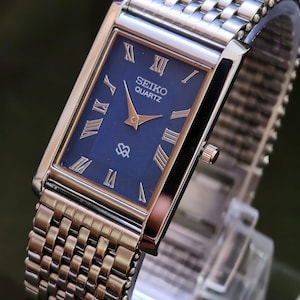 May include: A rectangular Seiko Quartz watch with a blue face and gold hands and Roman numerals. The watch has a silver-tone metal case and a matching mesh-style strap. The watch is displayed on a clear stand.