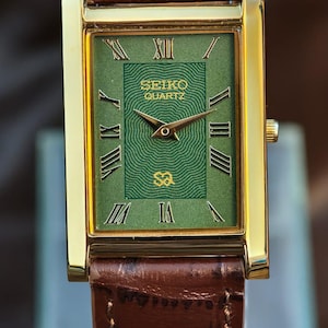 Może przedstawiać: Prostokątny, złoty zegarek Seiko Quartz z zieloną tarczą i brązowym sk&oacute;rzanym paskiem. Tarcza zegarka posiada rzymskie cyfry i logo Seiko. Brązowy sk&oacute;rzany pasek ma przeszycia.