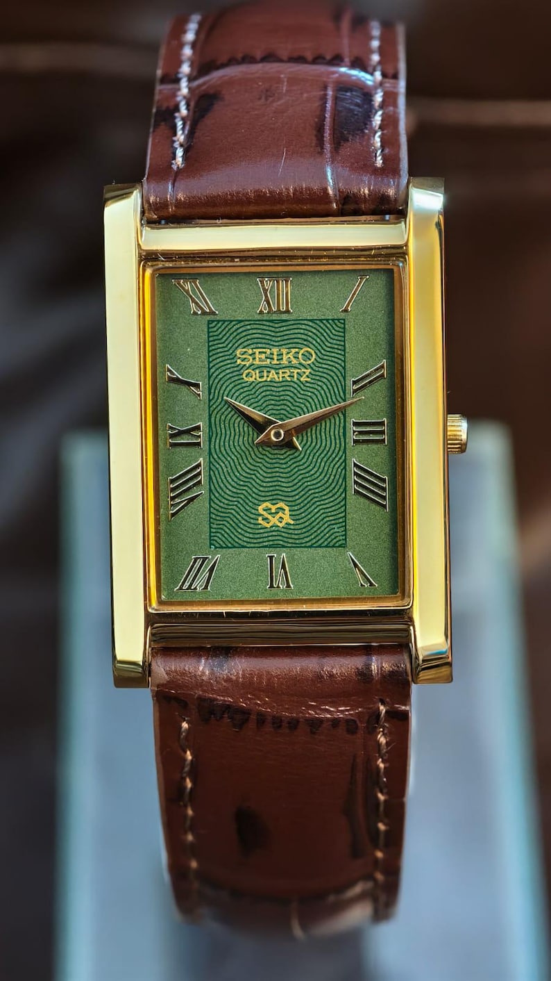 Może przedstawiać: Prostokątny, złoty zegarek Seiko Quartz z zieloną tarczą i brązowym sk&oacute;rzanym paskiem. Tarcza zegarka posiada rzymskie cyfry i logo Seiko. Brązowy sk&oacute;rzany pasek ma przeszycia.