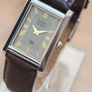 May include: A rectangular Seiko Quartz watch with a silver case and a brown leather band. The watch face is gray with gold Roman numerals and hands. The band is a dark brown with a textured pattern.