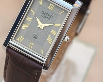 May include: A rectangular Seiko Quartz watch with a silver case and a brown leather band. The watch face is gray with gold Roman numerals and hands. The band is a dark brown with a textured pattern.
