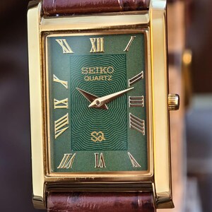 Może przedstawiać: Prostokątny, złoty zegarek Seiko Quartz z zieloną tarczą i złotymi rzymskimi cyframi. Zegarek ma brązowy sk&oacute;rzany pasek. Tarcza zegarka zawiera logo Seiko Quartz i złote wskaz&oacute;wki.