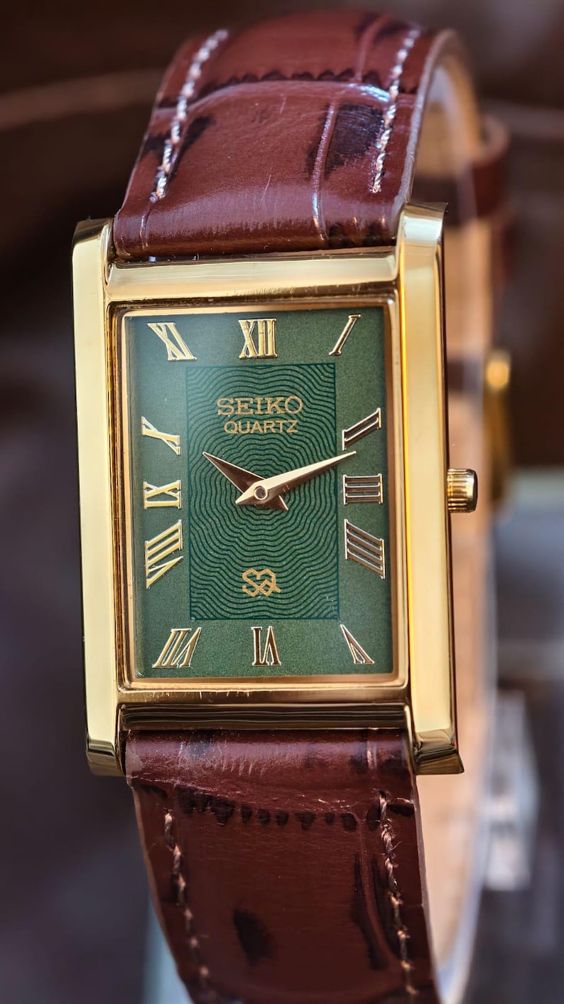Może przedstawiać: Prostokątny, złoty zegarek Seiko Quartz z zieloną tarczą i złotymi rzymskimi cyframi. Zegarek ma brązowy sk&oacute;rzany pasek. Tarcza zegarka zawiera logo Seiko Quartz i złote wskaz&oacute;wki.