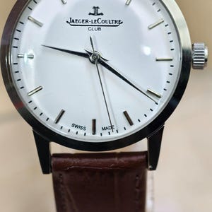 May include: A silver-toned Jaeger-LeCoultre Club watch with a white face, black hands, and a brown leather strap. The watch face features the Jaeger-LeCoultre logo and the words "SWISS MADE". The watch has a round face and a textured crown.