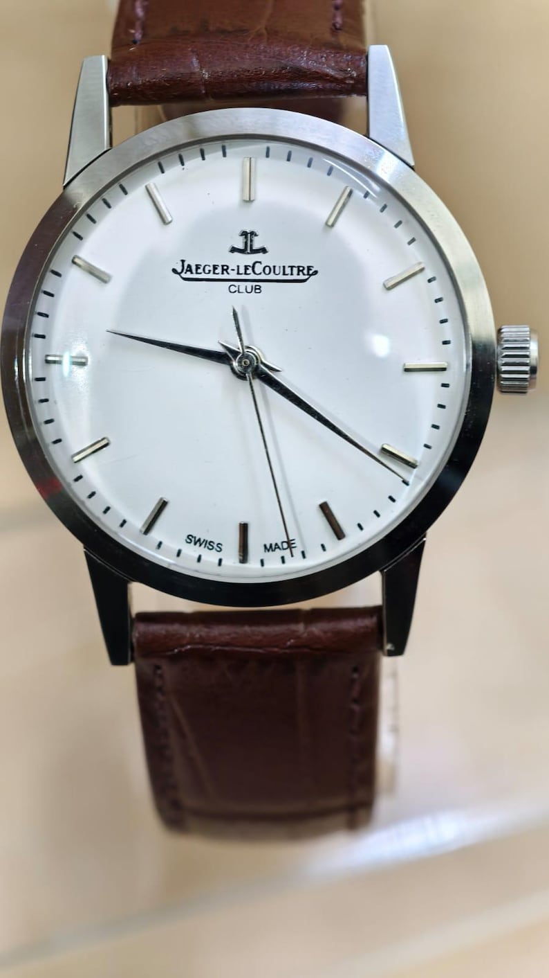 May include: A silver-toned Jaeger-LeCoultre Club watch with a white face, black hands, and a brown leather strap. The watch face features the Jaeger-LeCoultre logo and the words "SWISS MADE". The watch has a round face and a textured crown.