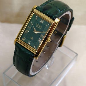 May include: A rectangular Seiko Quartz watch with a green face and gold-colored frame. The watch has a green leather band. Roman numerals mark the hours. The watch is displayed on a clear, rectangular stand.