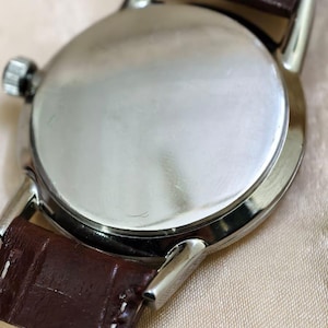 May include: Close-up of a wristwatch with a silver-colored case and a brown leather strap. The watch has a round face and a textured leather band. The watch is a classic style.