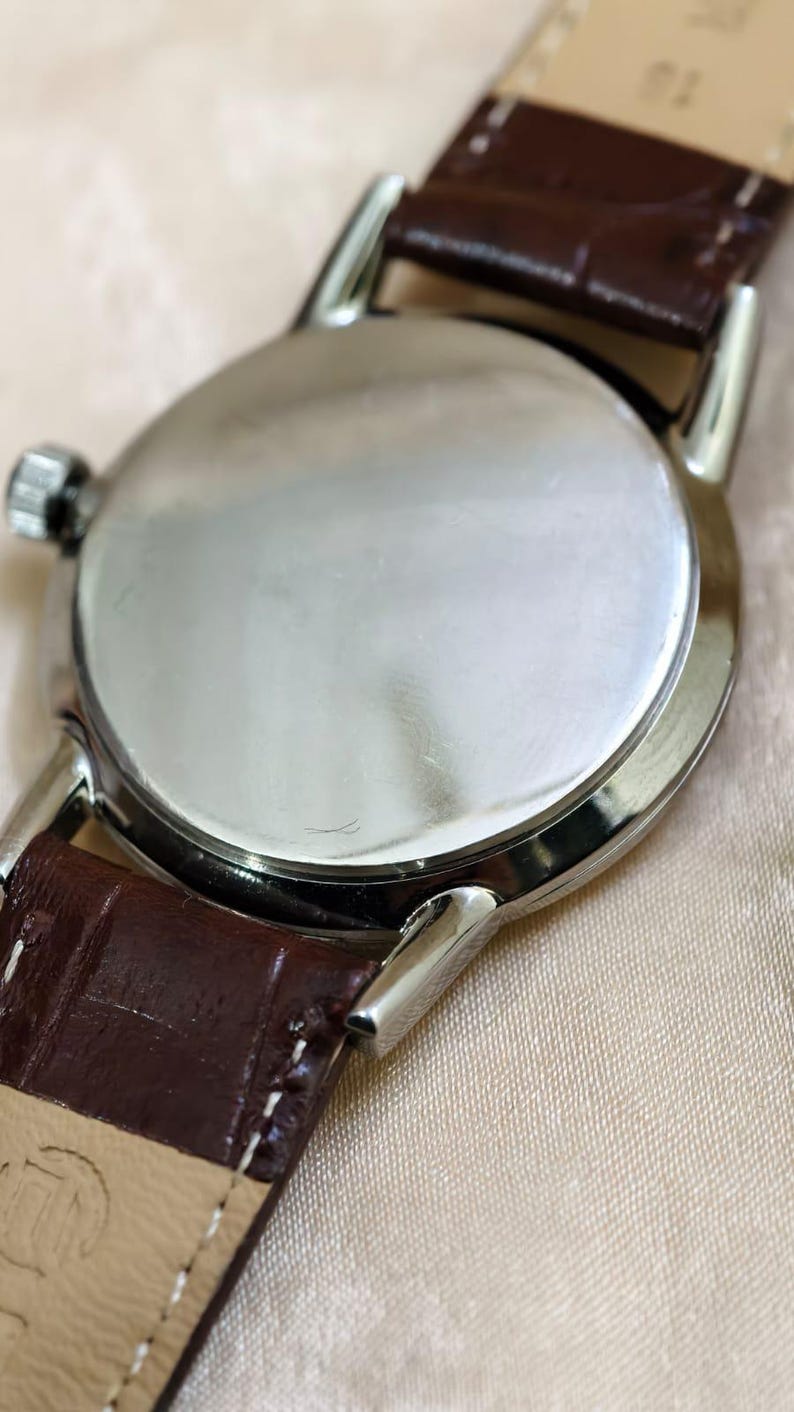 May include: Close-up of a wristwatch with a silver-colored case and a brown leather strap. The watch has a round face and a textured leather band. The watch is a classic style.