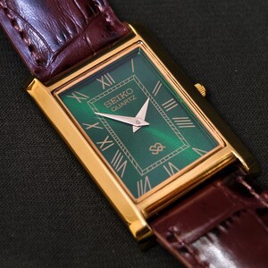 May include: A rectangular gold-toned Seiko Quartz watch with a green face and Roman numeral hour markers. The watch has gold hands and a brown leather band with a crocodile pattern.