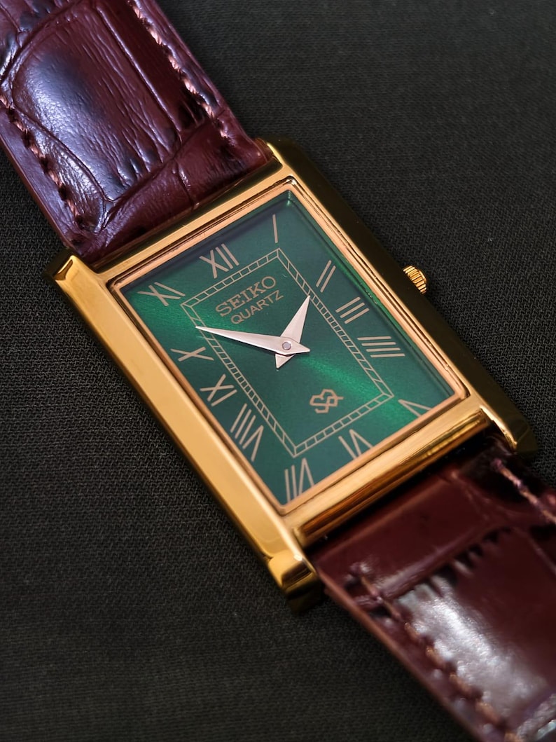 May include: A rectangular gold-toned Seiko Quartz watch with a green face and Roman numeral hour markers. The watch has gold hands and a brown leather band with a crocodile pattern.