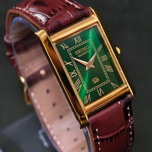 May include: A rectangular gold-toned Seiko Quartz watch with a green face and gold Roman numerals. The watch has a burgundy leather band. The watch face displays the Seiko Quartz logo. The watch band is approximately 8 inches long.