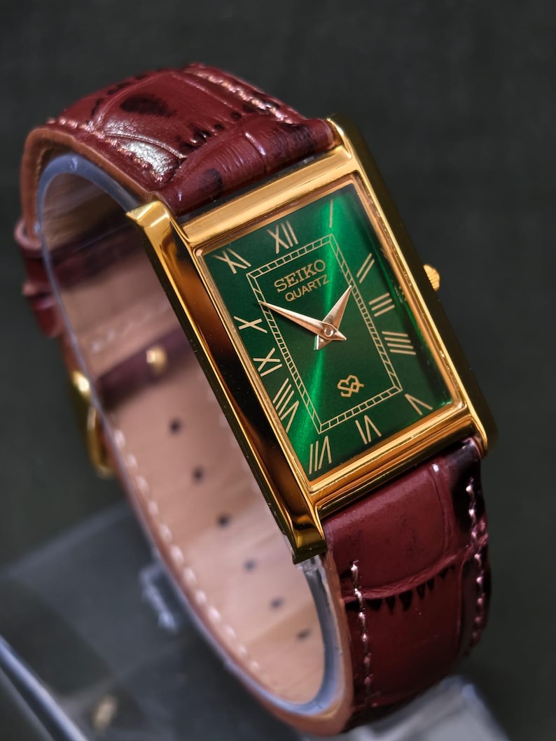 May include: A rectangular gold-toned Seiko Quartz watch with a green face and gold Roman numerals. The watch has a burgundy leather band. The watch face displays the Seiko Quartz logo. The watch band is approximately 8 inches long.