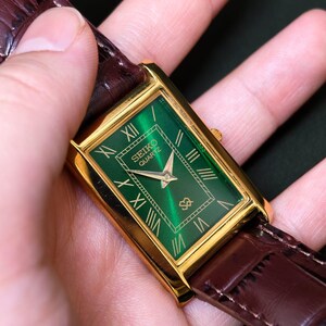 May include: A gold-toned rectangular Seiko Quartz watch with a green face and gold Roman numeral hour markers. The watch has a brown leather band.