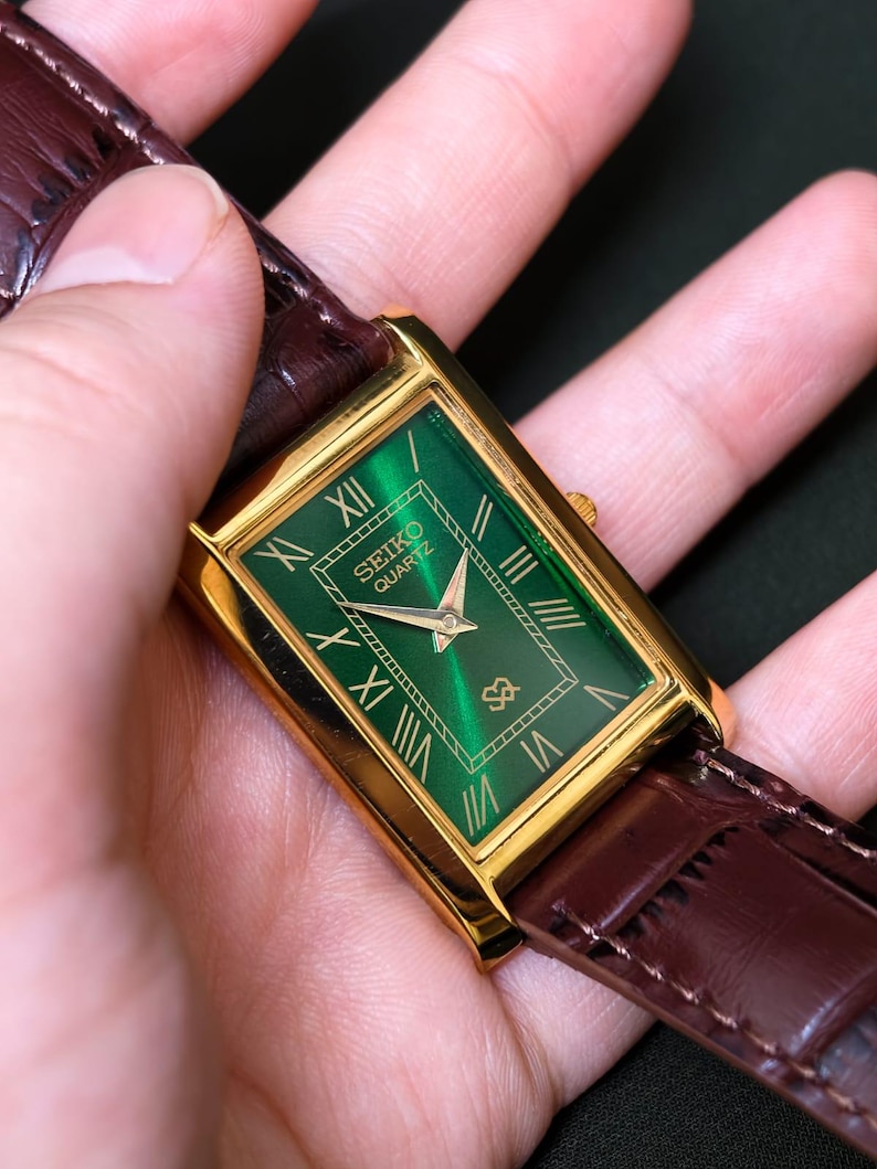 May include: A gold-toned rectangular Seiko Quartz watch with a green face and gold Roman numeral hour markers. The watch has a brown leather band.