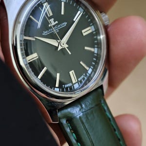 May include: A silver-cased wristwatch with a dark green face and a green leather band. The watch face features silver hands and markers, with the brand name "Jaeger-LeCoultre" visible. The watch band is a textured green leather.