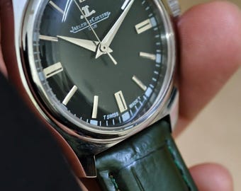 Può includere: Un orologio da polso argentato con quadrante verde scuro e cinturino in pelle verde. Il quadrante dell'orologio presenta lancette e indicatori argentati, con il marchio "Jaeger-LeCoultre" visibile. Il cinturino è in pelle verde strutturata.