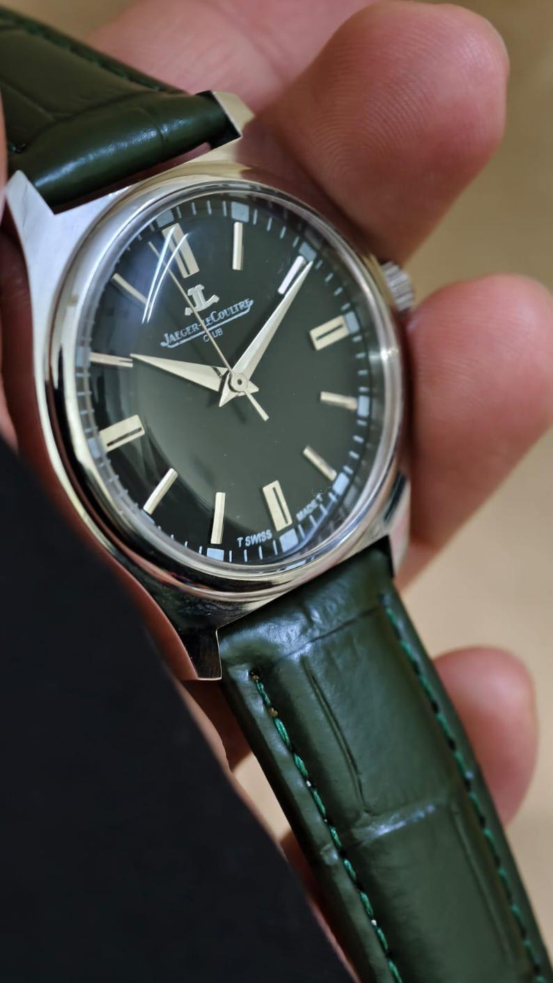 May include: A silver-cased wristwatch with a dark green face and a green leather band. The watch face features silver hands and markers, with the brand name "Jaeger-LeCoultre" visible. The watch band is a textured green leather.