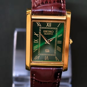 May include: A gold-toned rectangular Seiko Quartz watch with a green face and Roman numeral hour markers. The watch has gold hands and a brown leather band. The watch face has the Seiko Quartz logo.