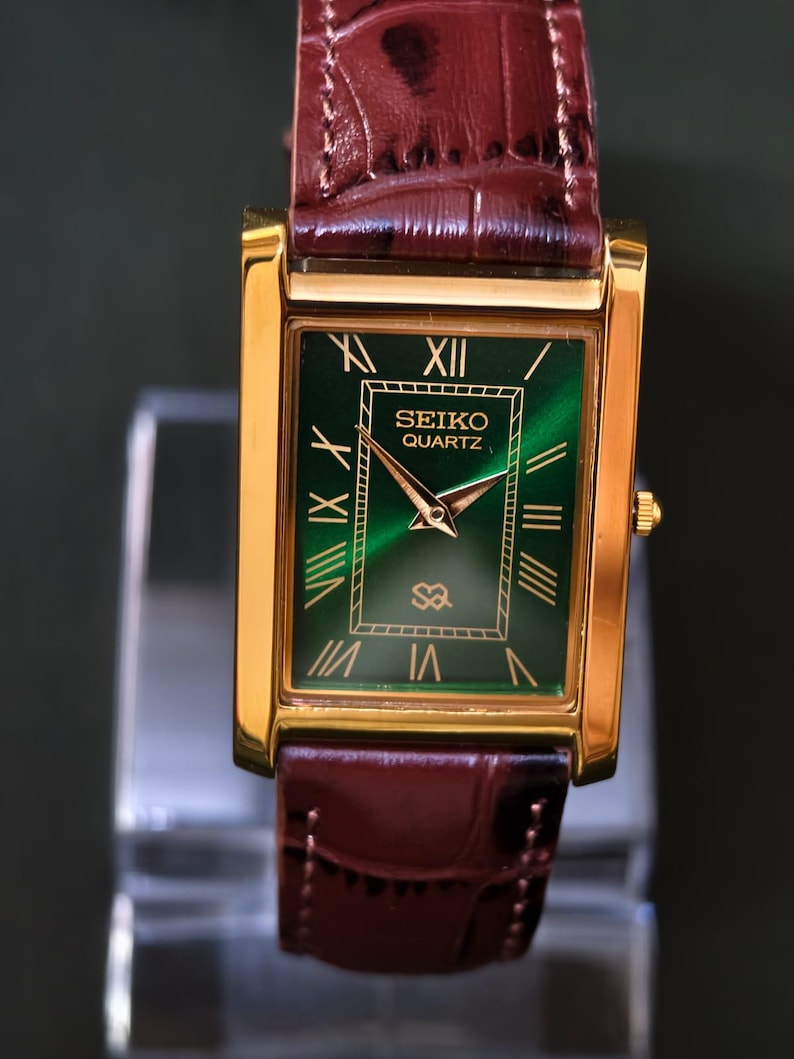 May include: A gold-toned rectangular Seiko Quartz watch with a green face and Roman numeral hour markers. The watch has gold hands and a brown leather band. The watch face has the Seiko Quartz logo.