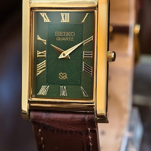 Może przedstawiać: Złoty zegarek Seiko Quartz z prostokątną zieloną tarczą i rzymskimi cyframi. Zegarek ma złote wskaz&oacute;wki i brązowy sk&oacute;rzany pasek. Na tarczy widnieje logo Seiko Quartz.