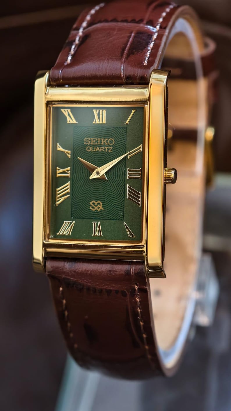 Może przedstawiać: Złoty zegarek Seiko Quartz z prostokątną zieloną tarczą i rzymskimi cyframi. Zegarek ma złote wskaz&oacute;wki i brązowy sk&oacute;rzany pasek. Na tarczy widnieje logo Seiko Quartz.