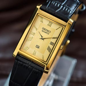 May include: A gold-toned Seiko Quartz watch with a rectangular face and black leather band. The watch face features Roman numerals and a textured center. The watch is displayed on a clear stand.