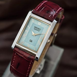 May include: A rectangular Seiko Quartz watch with a light blue face and gold-tone hands and accents. The watch has a silver-tone case and a burgundy leather band. The watch face displays Roman numerals and the Seiko Quartz logo.