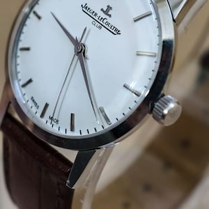 May include: A silver-toned Jaeger-LeCoultre Club watch with a white face and brown leather strap. The watch face features black hour markers and the words "JAEGER-LECOULTRE CLUB SWISS MADE". The watch has a classic design.