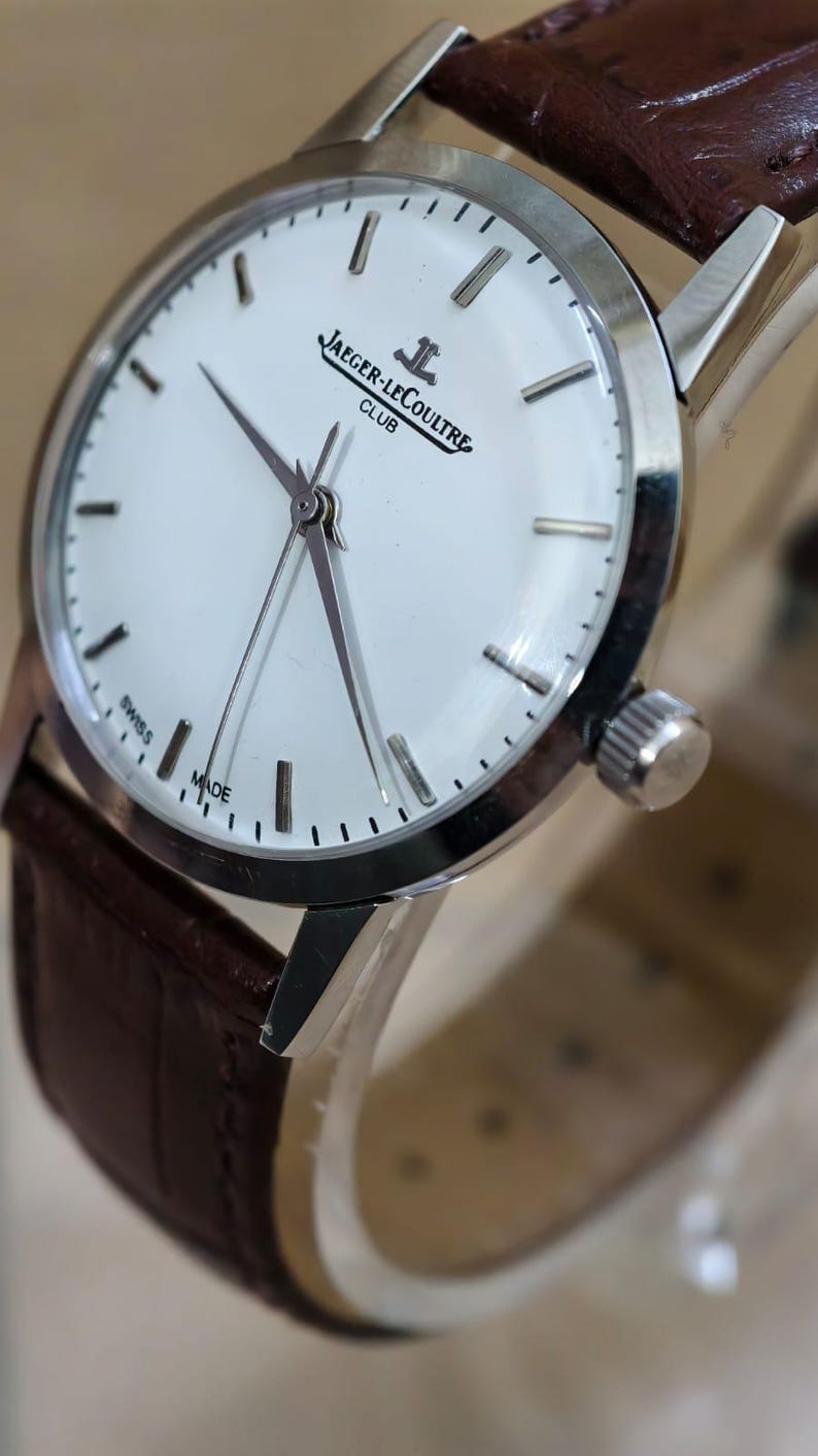 May include: A silver-toned Jaeger-LeCoultre Club watch with a white face and brown leather strap. The watch face features black hour markers and the words "JAEGER-LECOULTRE CLUB SWISS MADE". The watch has a classic design.