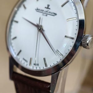 May include: A silver-toned wristwatch with a white face and brown leather band. The watch features a minimalist design with silver hour markers and hands. The watch face displays the brand name "Jaeger-LeCoultre" and the word "CLUB".