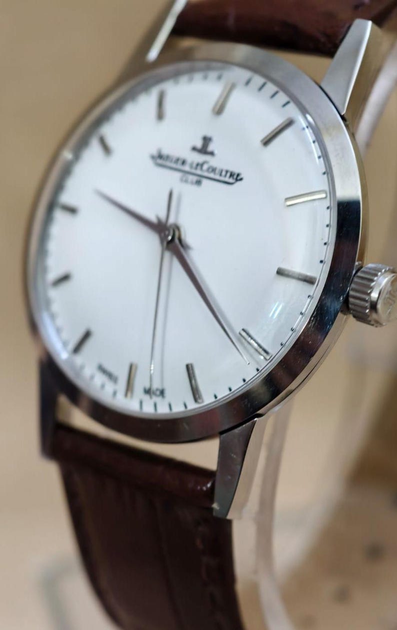 May include: A silver-toned wristwatch with a white face and brown leather band. The watch features a minimalist design with silver hour markers and hands. The watch face displays the brand name "Jaeger-LeCoultre" and the word "CLUB".