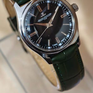 May include: A silver-toned wristwatch with a black face and a dark green leather band. The watch face features silver hands and hour markers. The watch is branded with the text "Jaeger-LeCoultre Club" and "T Swiss".