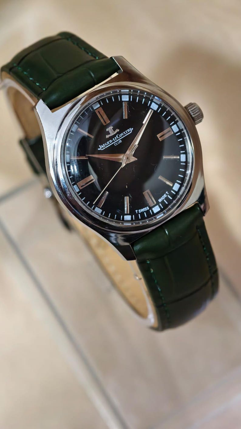 May include: A silver-toned wristwatch with a black face and a dark green leather band. The watch face features silver hands and hour markers. The watch is branded with the text "Jaeger-LeCoultre Club" and "T Swiss".