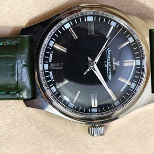 May include: A silver-toned wristwatch with a black face and a green leather band. The watch face features silver-toned hands and markers, with the brand name "Jaeger-LeCoultre Club" visible. The green band is approximately 0.75 inches wide.