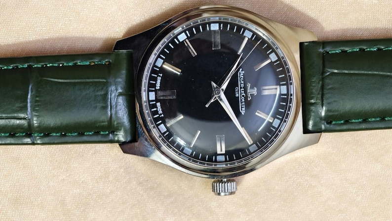 May include: A silver-toned wristwatch with a black face and a green leather band. The watch face features silver-toned hands and markers, with the brand name "Jaeger-LeCoultre Club" visible. The green band is approximately 0.75 inches wide.