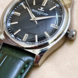 May include: Close-up of a Jaeger-LeCoultre Club watch with a dark dial and silver case. The watch features silver hands and markers, and a green leather strap. The watch face displays the Jaeger-LeCoultre logo and the words "CLUB" and "T SWISS MADE T".