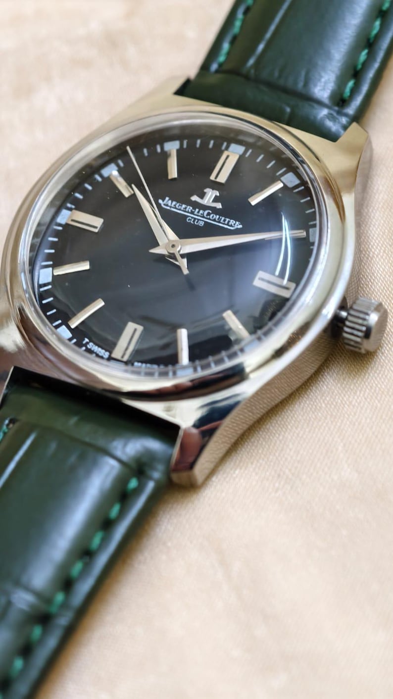May include: Close-up of a Jaeger-LeCoultre Club watch with a dark dial and silver case. The watch features silver hands and markers, and a green leather strap. The watch face displays the Jaeger-LeCoultre logo and the words "CLUB" and "T SWISS MADE T".