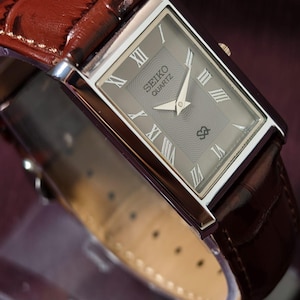 Puede incluir: Un reloj Seiko Quartz rectangular con caja plateada y esfera gris con números romanos. El reloj tiene una correa de cuero marrón con un patrón de cocodrilo.