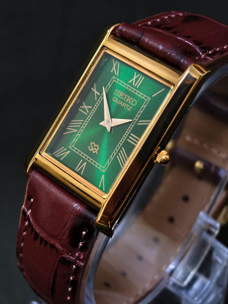May include: A rectangular Seiko Quartz watch with a gold-tone case and a green dial. The dial features gold Roman numerals and hands. The watch has a brown leather band with a crocodile pattern.