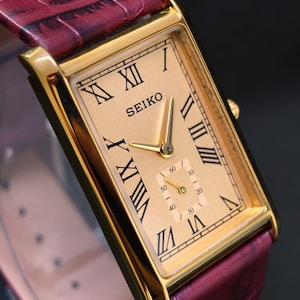Könnte beinhalten: Goldfarbene Seiko-Uhr mit rechteckigem Zifferblatt und römischen Ziffern. Die Uhr hat ein kleines Sekunden-Zifferblatt und ein bordeauxfarbenes Lederarmband. Das Zifferblatt ist ca. 3 cm breit.