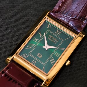 May include: A rectangular gold-toned Seiko Quartz watch with a green face and Roman numeral hour markers. The watch has a brown leather band. The watch face displays the Seiko Quartz logo.