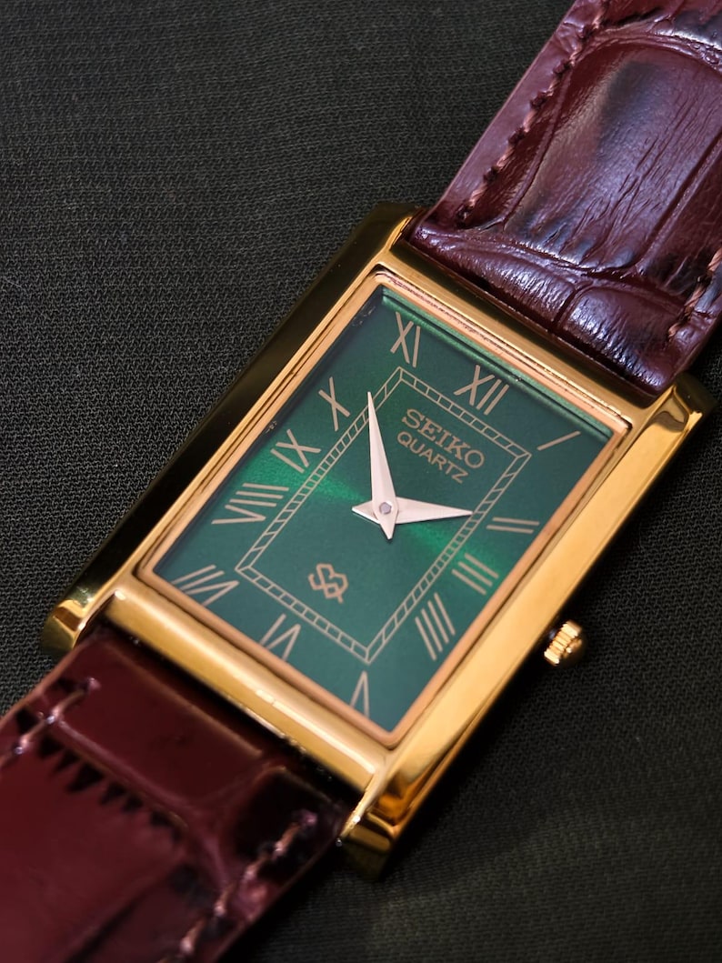 May include: A rectangular gold-toned Seiko Quartz watch with a green face and Roman numeral hour markers. The watch has a brown leather band. The watch face displays the Seiko Quartz logo.